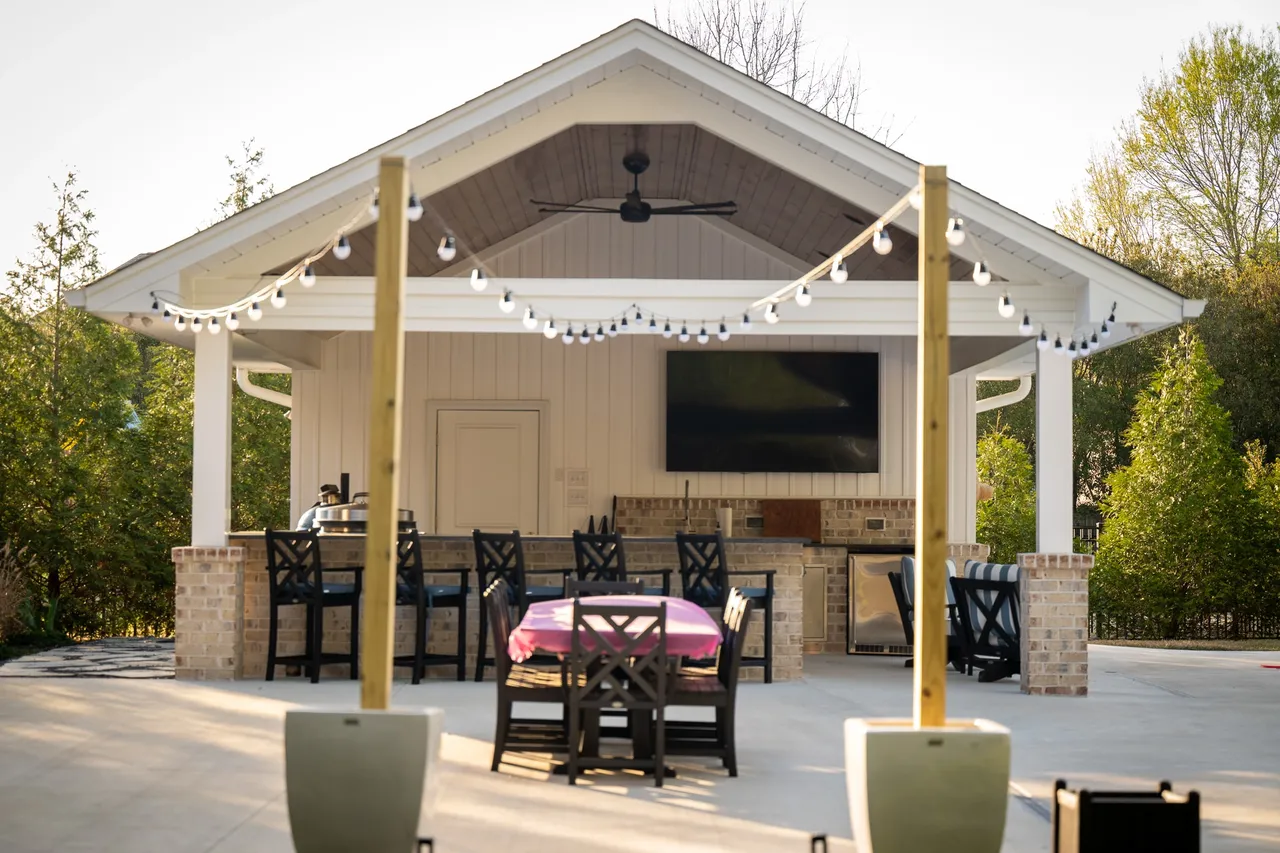 White pavilion with brick bar, mounted TV, string lights, and dining set at sunset