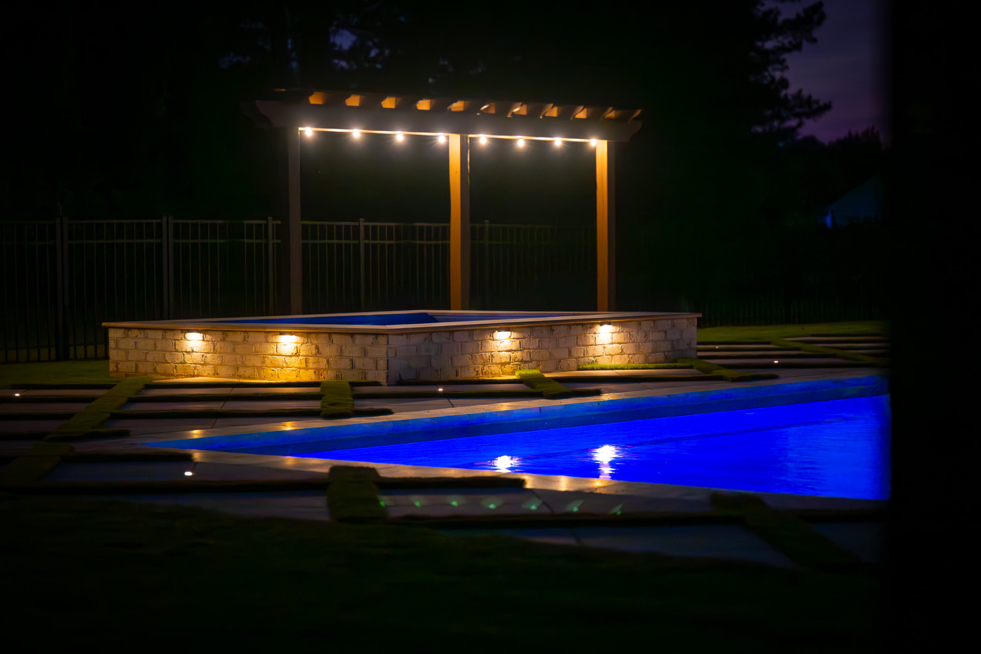 Raised brick spa beneath a lit pergola beside a blue-lit pool at night