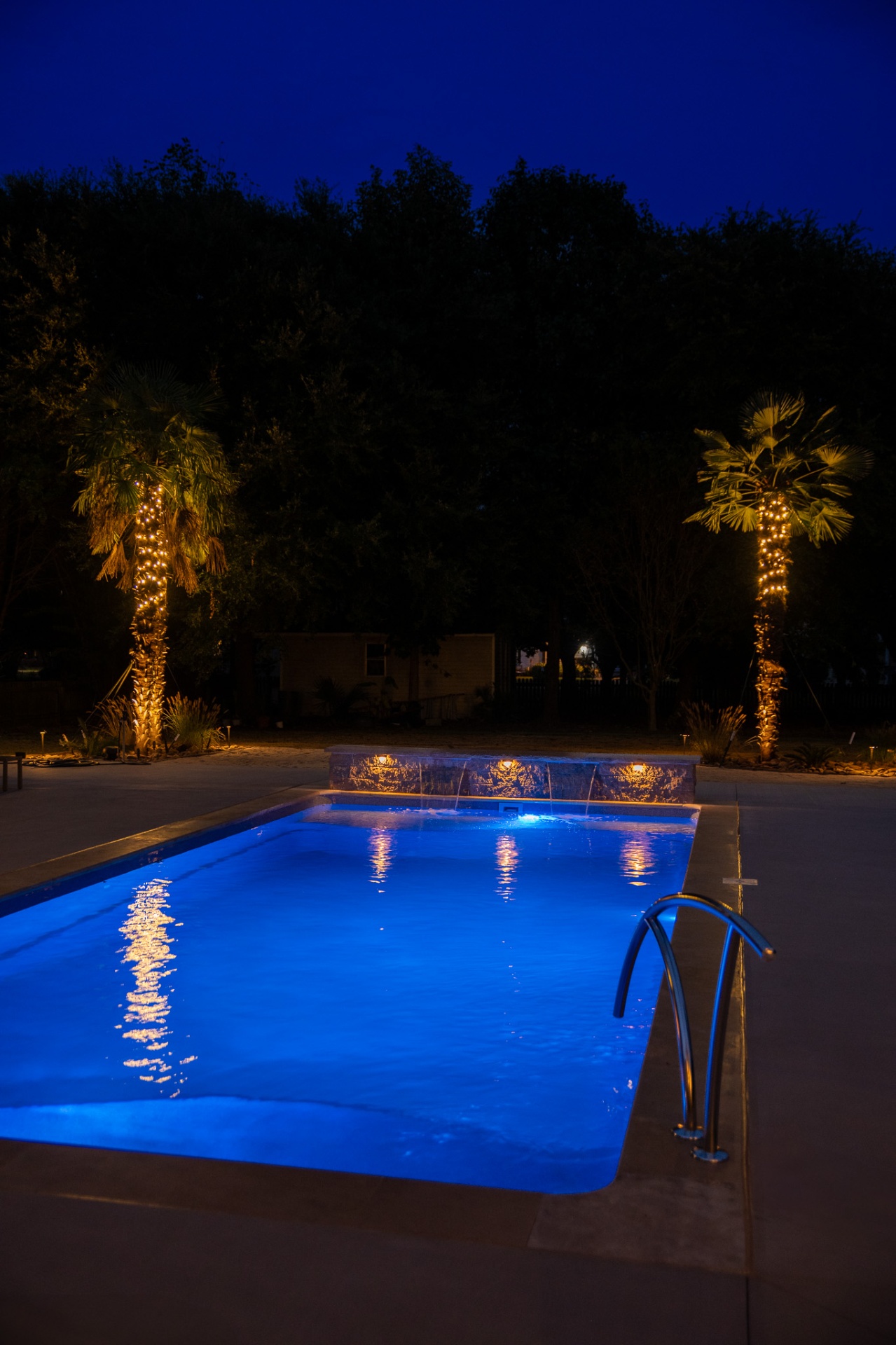 Pool at dusk framed by lit palm trees and stacked stone waterfall