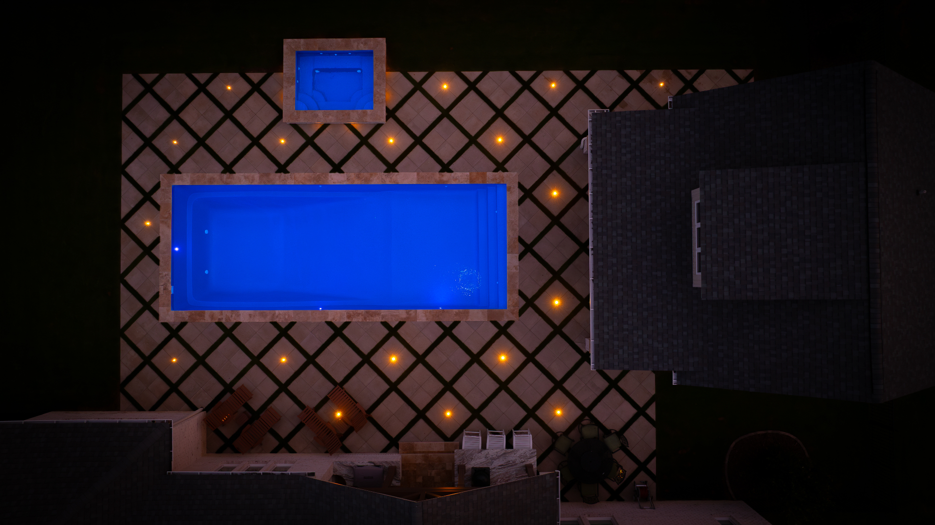 Overhead night view of a rectangular pool and spa illuminated blue with warm deck lights on a diamond paver pattern