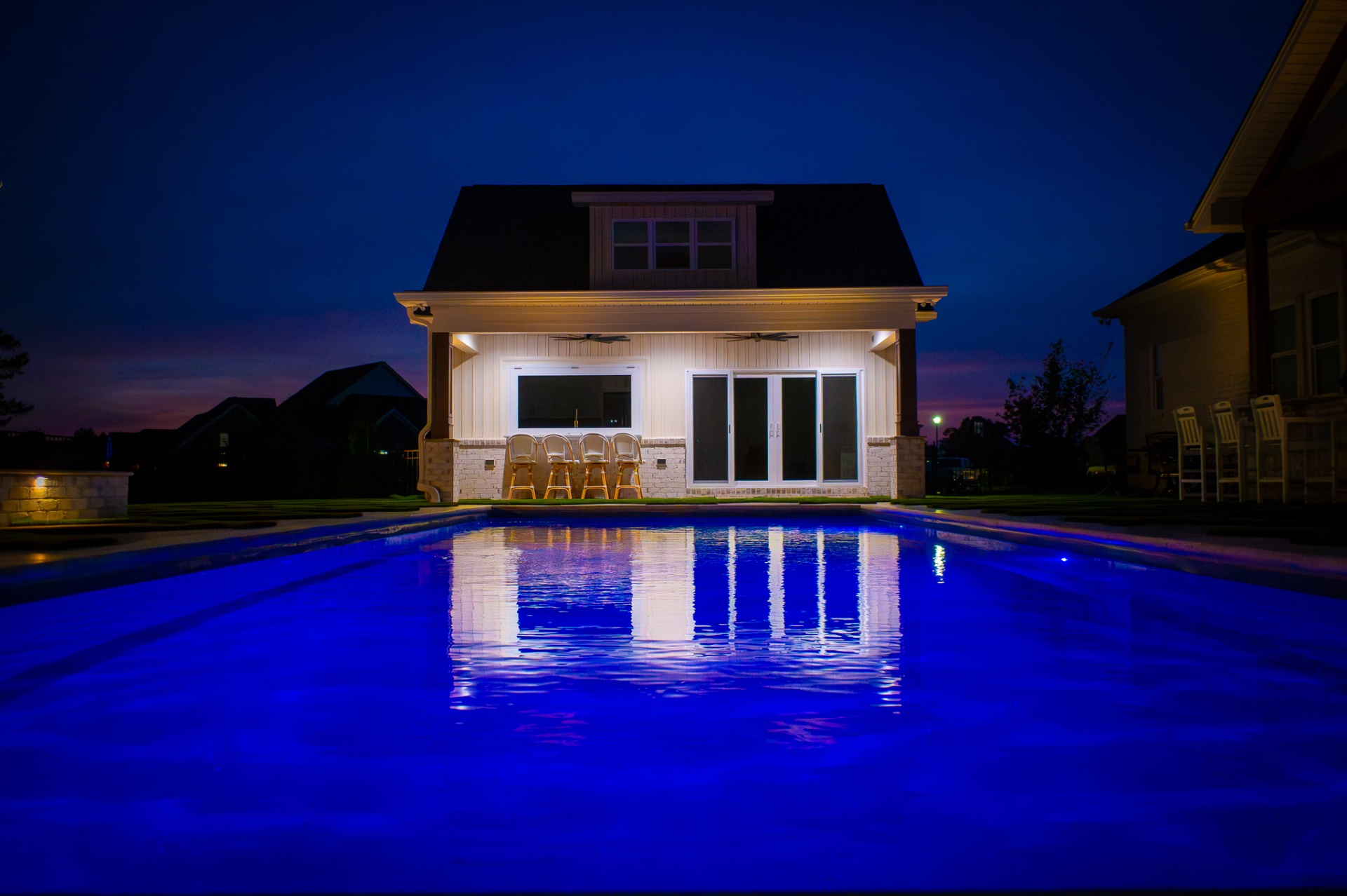 Pool house at dusk with warm covered patio lighting reflecting on a blue-lit pool