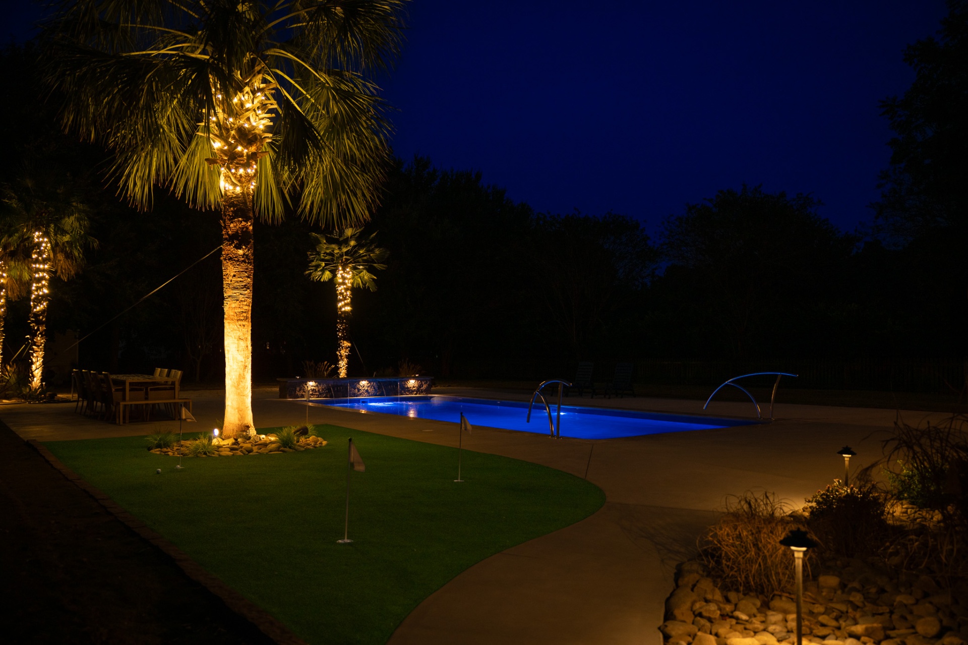 Backyard at night with lit palms, putting green, and glowing blue pool